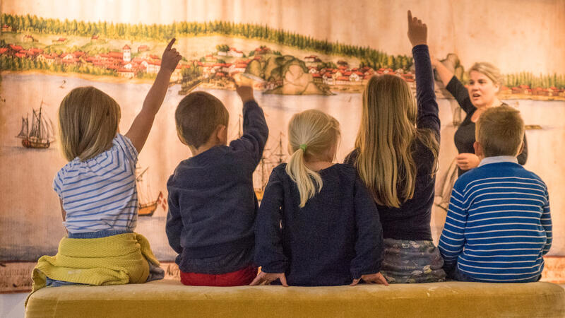 Fem barn følger med på en formidler som forteller. Flere av barna rekker opp hånden. I bakgrunnen ser vi et gammelt bilde av Larvik by.