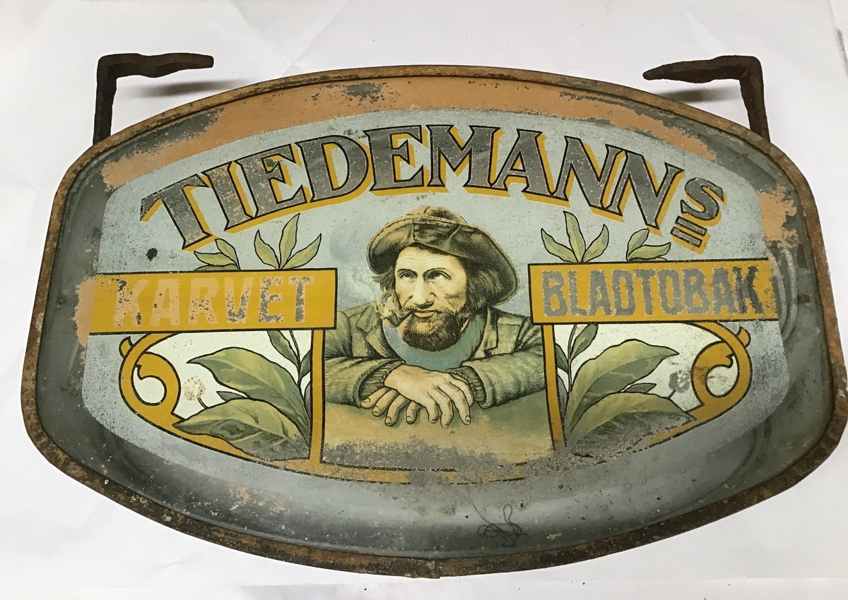 Reklameskilt for Tiedemanns tobakksvarer. Skiltet er avlang, ellipseforma men med skrå kortsider. Ulike motiv på sidene, det er opplegg for elektrisk lys med lyspærer  inne i skiltet.