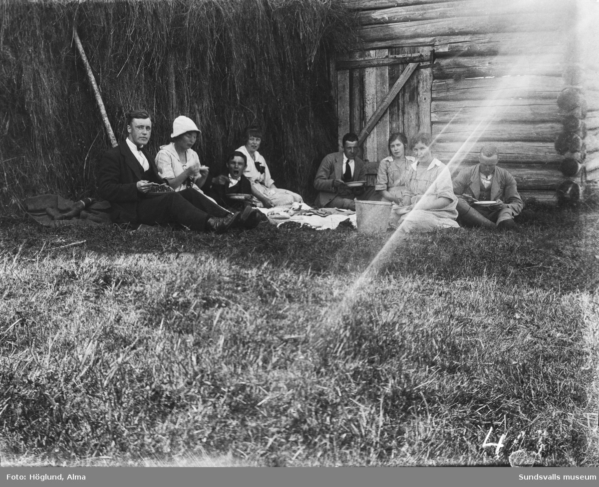 Fäbodbild med en grupp män och kvinnor som sitter i gräset och äter.