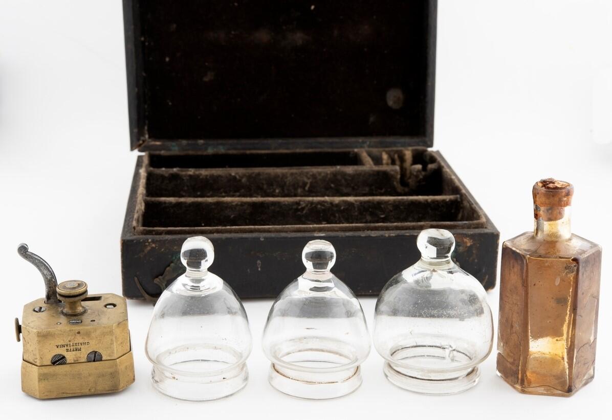 bildet viser en liten mørk kasse med små rom og åpent lokk. Foran ser vi en messingdings, tre glasskupler i klart glass og en brun glassflaske.