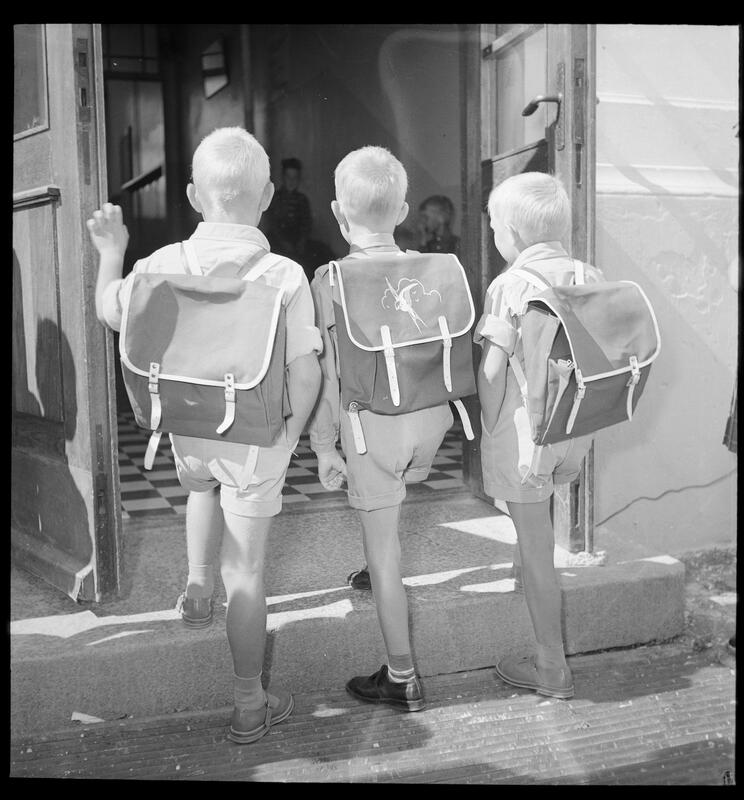 Sort-hvit-bildet viser ryggene til tre små gutter i shorts og kortermet genser og helt like skolesekker. De går opp et trinn og inn en dør. (Foto/Photo)