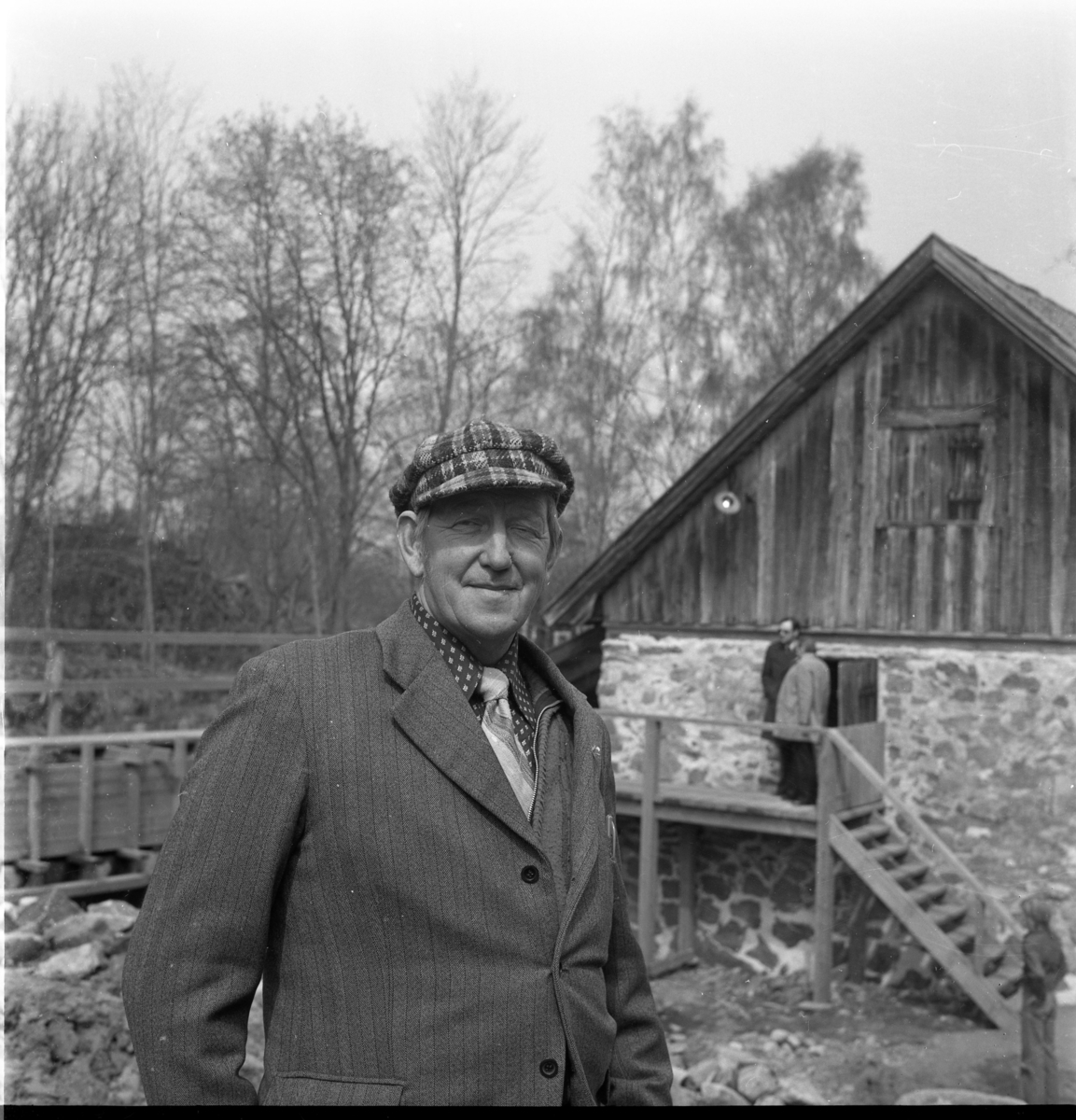Porträtt av en man med keps, slips och blazer stående vid Rasmus kvarn östra gavel.