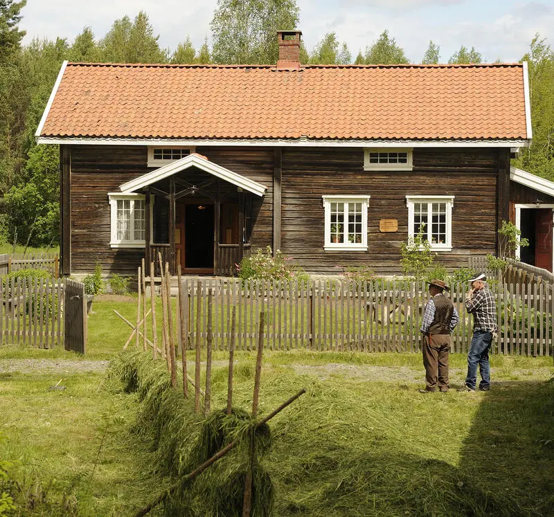 Storbråten på Odalstunet. Et tømmerhus med rødt tak. En etasje og et loft. Foran Storbråten-huset er det en inngjerdet hage. Foran hagen er det to menn. I forgrunn ser vi hesjing.