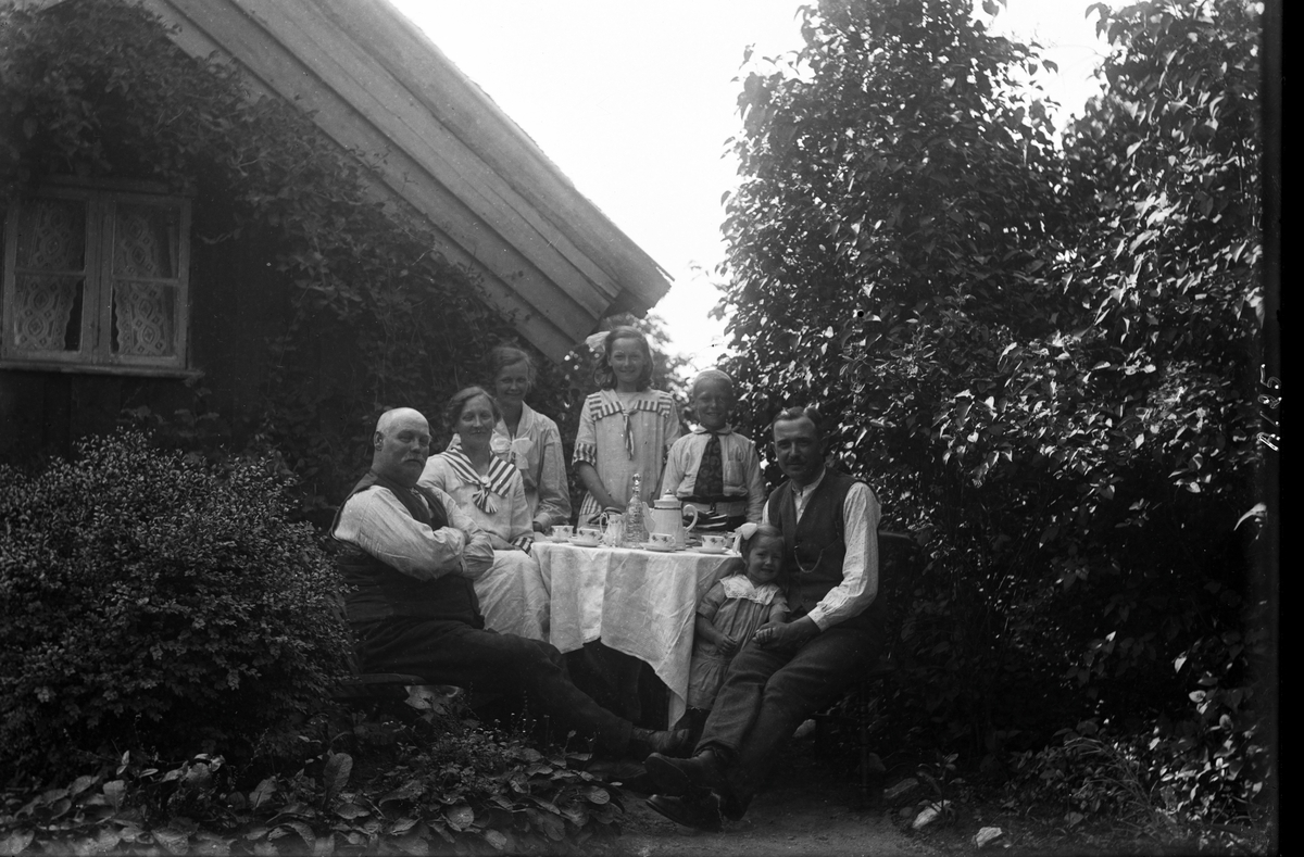 Vuxna och barn samlade kring ett kaffebord vid gaveln till en låg stuga, Lilla Gunnels i Sällstorp. Mannen till vänster heter Ahlgren. Uteplatsen är lummig, omgiven av höga syrenbuskar, en frodig klängväxt på husgaveln och annan växtlighet. Pojkens byxor hålls uppe av ett resårskärp, mannen till höger har en småmönstrad skjorta med ståkrage och ett par av klänningarna har detaljer av ett bredrandigt tyg. (Se även bild nr GEA132)