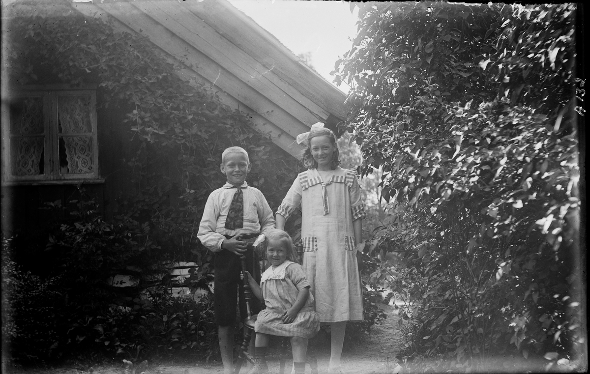 Tre barn, troligen syskon, vid gaveln till "Ahlgrens stuga", Lilla Gunnels i Sällstorp. Den minsta flickan sitter på en pinnstol och de andra står bakom henne. Till höger växer höga syrenbuskar och på husväggen klättrar klängväxter. Flickorna har rosetter i håret, pojken en bred slips och den äldre flickans klänning har detaljer av ett bredrandigt tyg. (Se även bild nr GEA125)