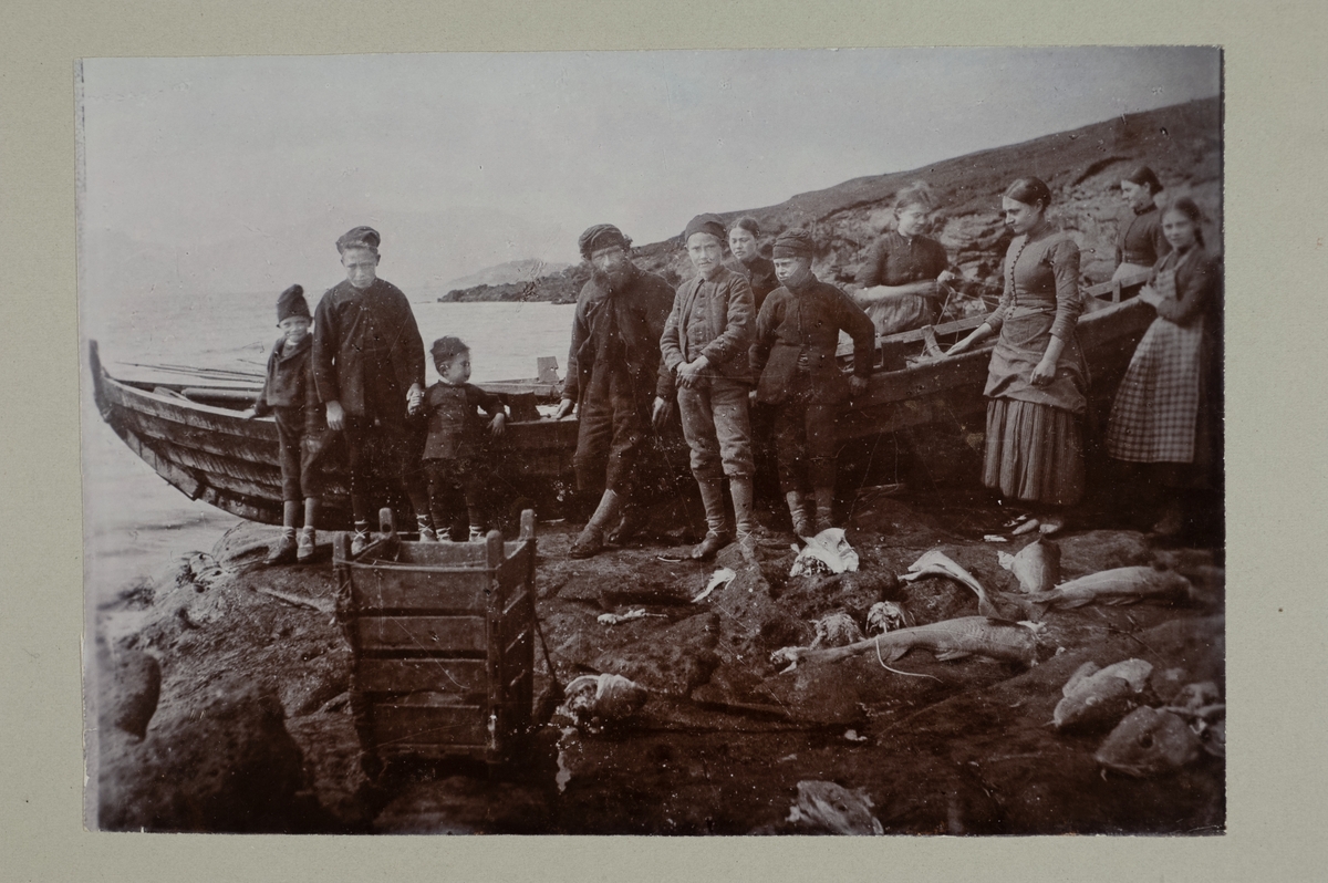 En grupp människor, vuxna och barn, vid en båt på en strand. Fisk ligger på marken framför dem. 
Anteckning på baksidan: "En hjemvendt Fiskeboud"