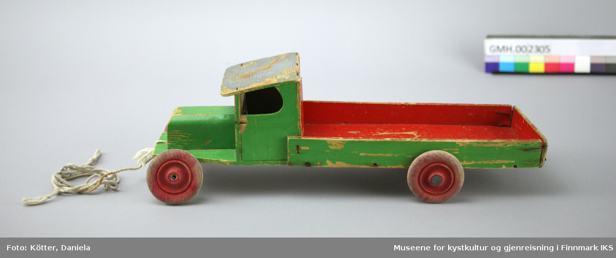 Lekebilen er laget av tre og kryssfinér. Den er malt grønn og rød. Bilen har en trekksnor festet i panseret
