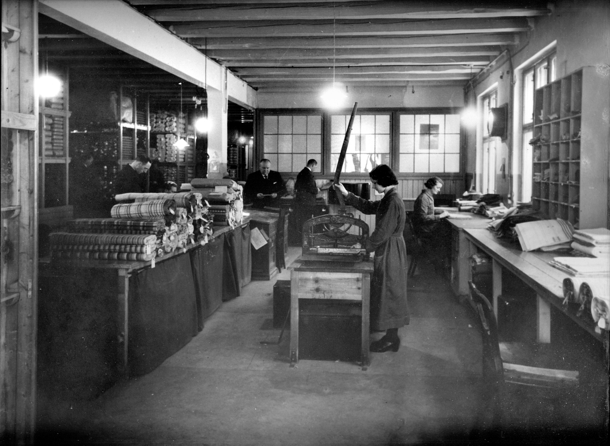 Produksjon av vareprøver på lageret til bomullsveveriet ved Knud Graah, kvinne ved skjæremaskinen, i tillegg en kvinne og fire menn i arbeid.
