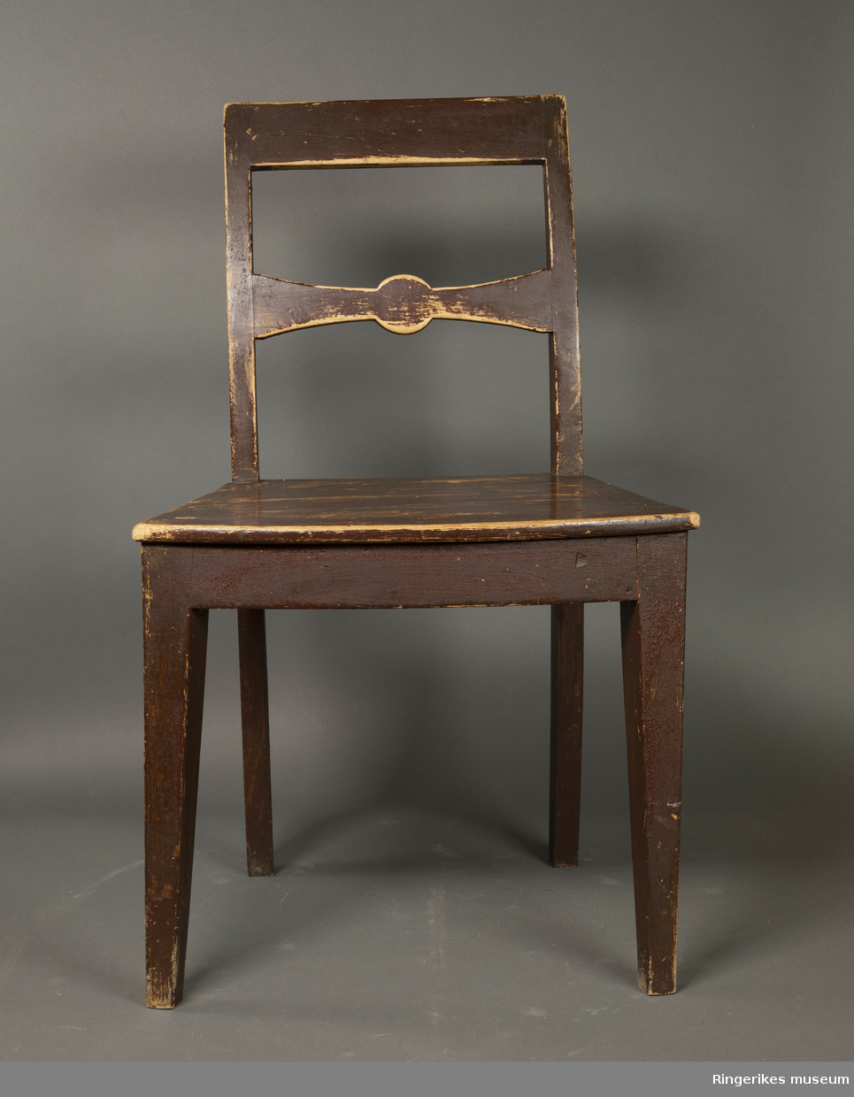 Brunmalt empire stol i furu. Stolen har ikke sprosser mellom stolbenene. Enkelt toppstykke og ryggbrett med sløyfeform. Sargen er bredere i front enn bak og har en svak bue i front.