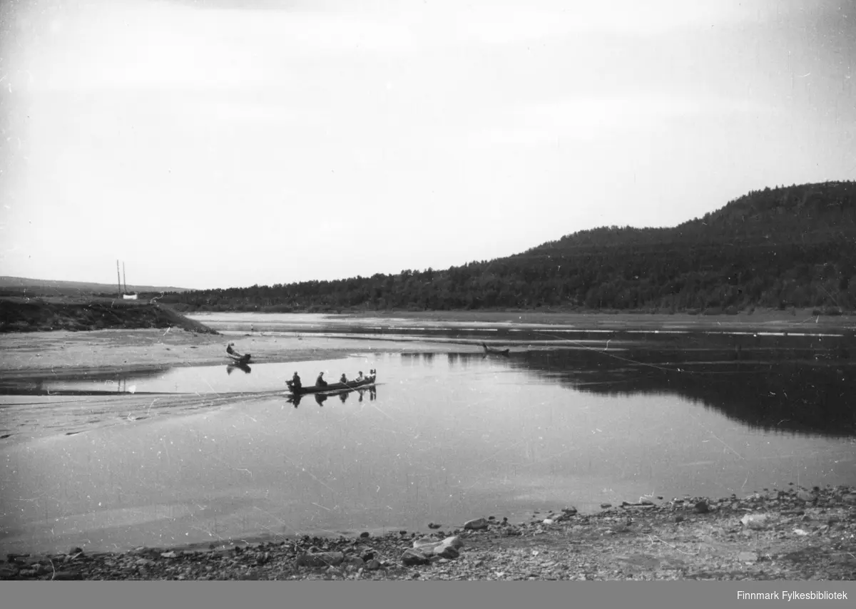 Med elvebåt på Karasjokka. En båt med fire personer kjører midt på elva. To elvebåter ligger ved elvebredden på andre siden av elva.