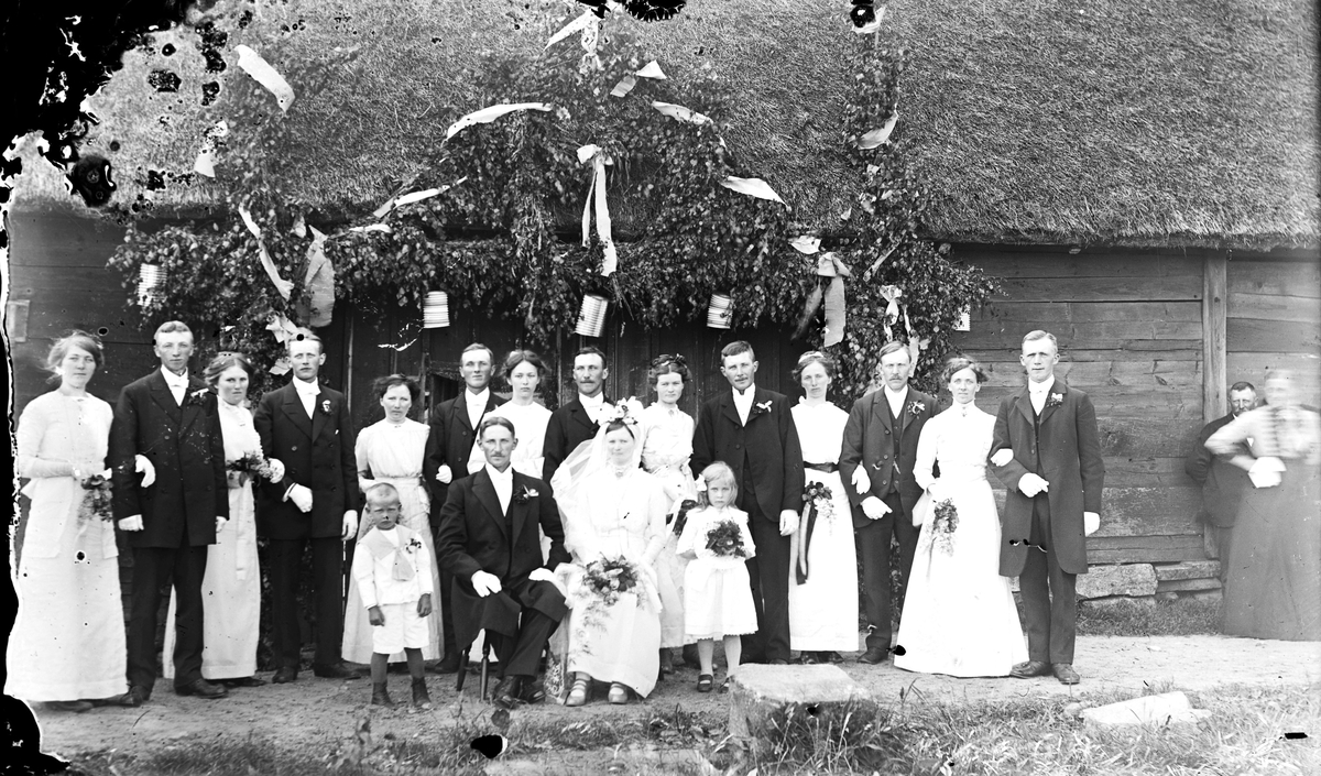 Svennerbergs bröllop, ca 1914 i Svenstorp, Larsa, Sällstorp. Brudparet sitter bredvid sina brudnäbbar och framför sina gäster, tärnor och marskalker. De är samlade vid en äreport med breda band och veckade pappersdekorer invid en byggnad av skiftesverk med halmtak. Längst till höger ses fotografens mor Bertha Engström.