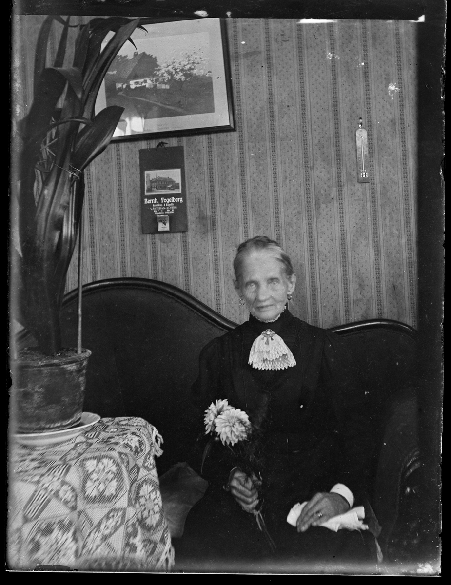 Äldre kvinna sitter i soffan med en blomma i handen.