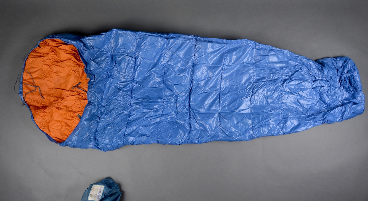 En sovepose i trekk/pose av merket Ajungilak (Nanok Babyble 4000). Soveposen har yttertrekk av blått kunststoff og er foret med oransje bomullsstoff. Soveposen er fylt med dun. Langs åpningen er det en løpekant der det er tredd i bomullsbånd for stramming. På snoren er det en enkel plastlås. På soveposen er det også snorer for å knyte rundt posen når den er rullet sammen. Til posen er det et blått nylontrekk med snøring i åpningen og en enkel plastlås for lukking. På soveposen og på trekket er det sydd på identiske merker, se "Påført tekst/merker".