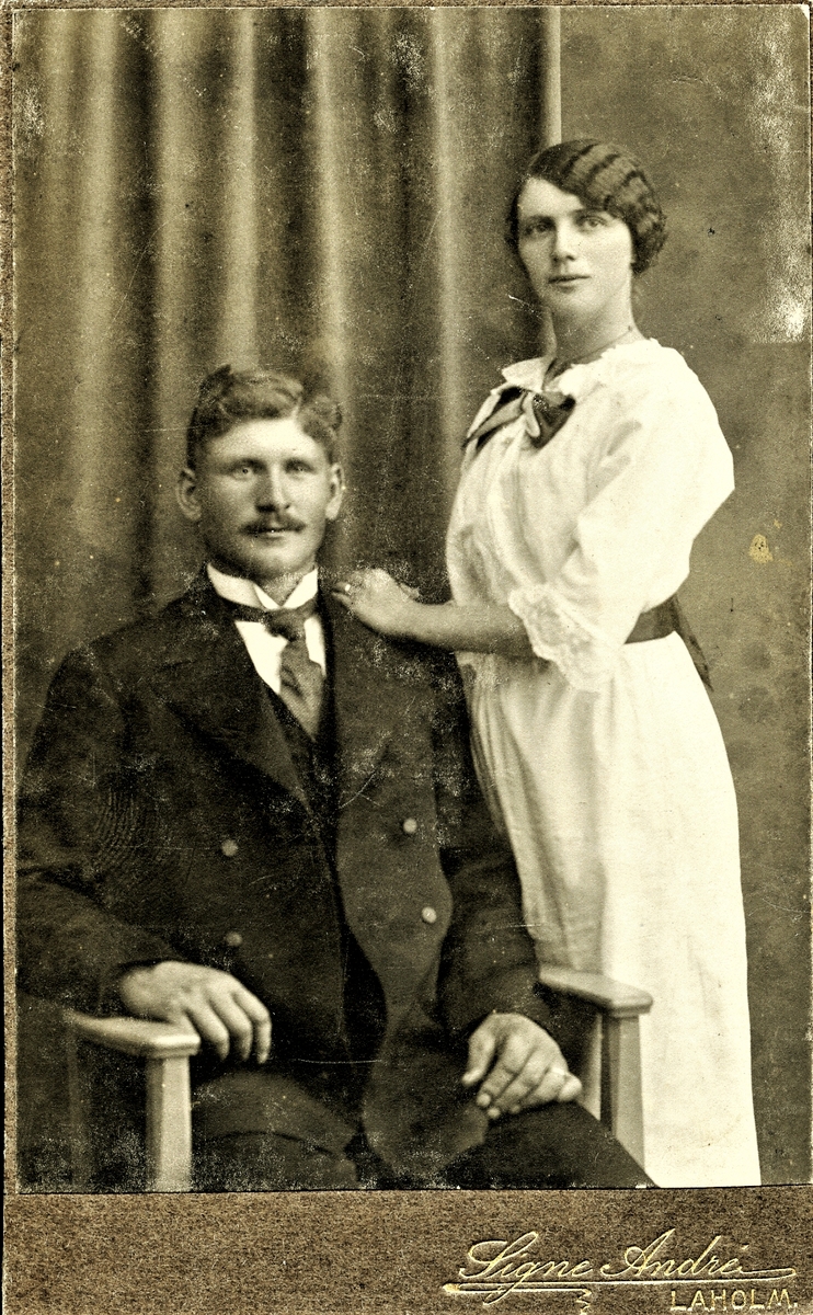Ett ungt, okänt par fotograferat i Laholm. Mannen sitter i en stol och kvinnan står bredvid med handen på hans axel. Båda bär ringar så troligen är det ett förlovningsfoto. hennes hår är uppsatt och ondulerat (vågigt). Han har sidbena och mustasch.