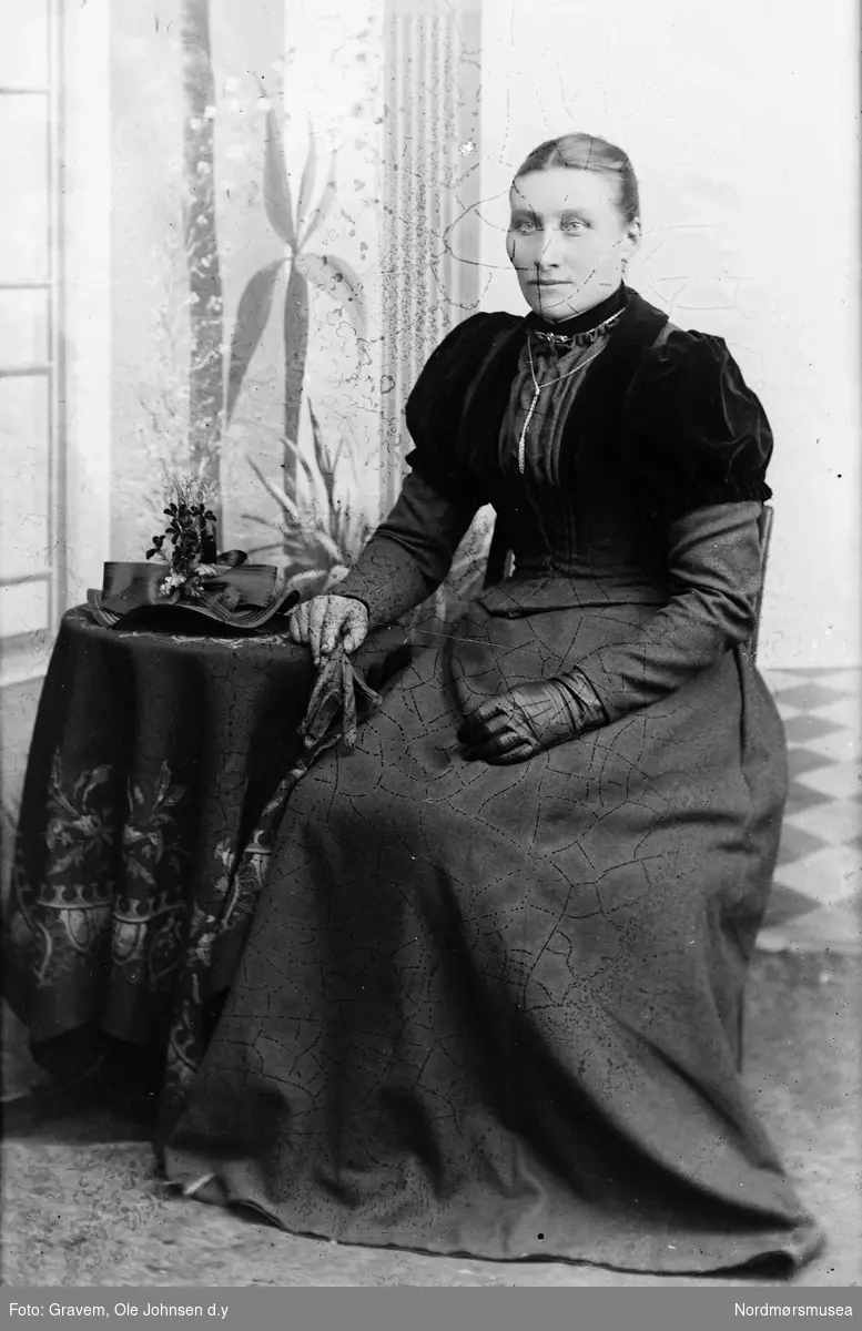 Portrett i helfigur av sittende kvinne som sitter ved et bord. Kvinnen har på seg kjole, hansker og smykke. 