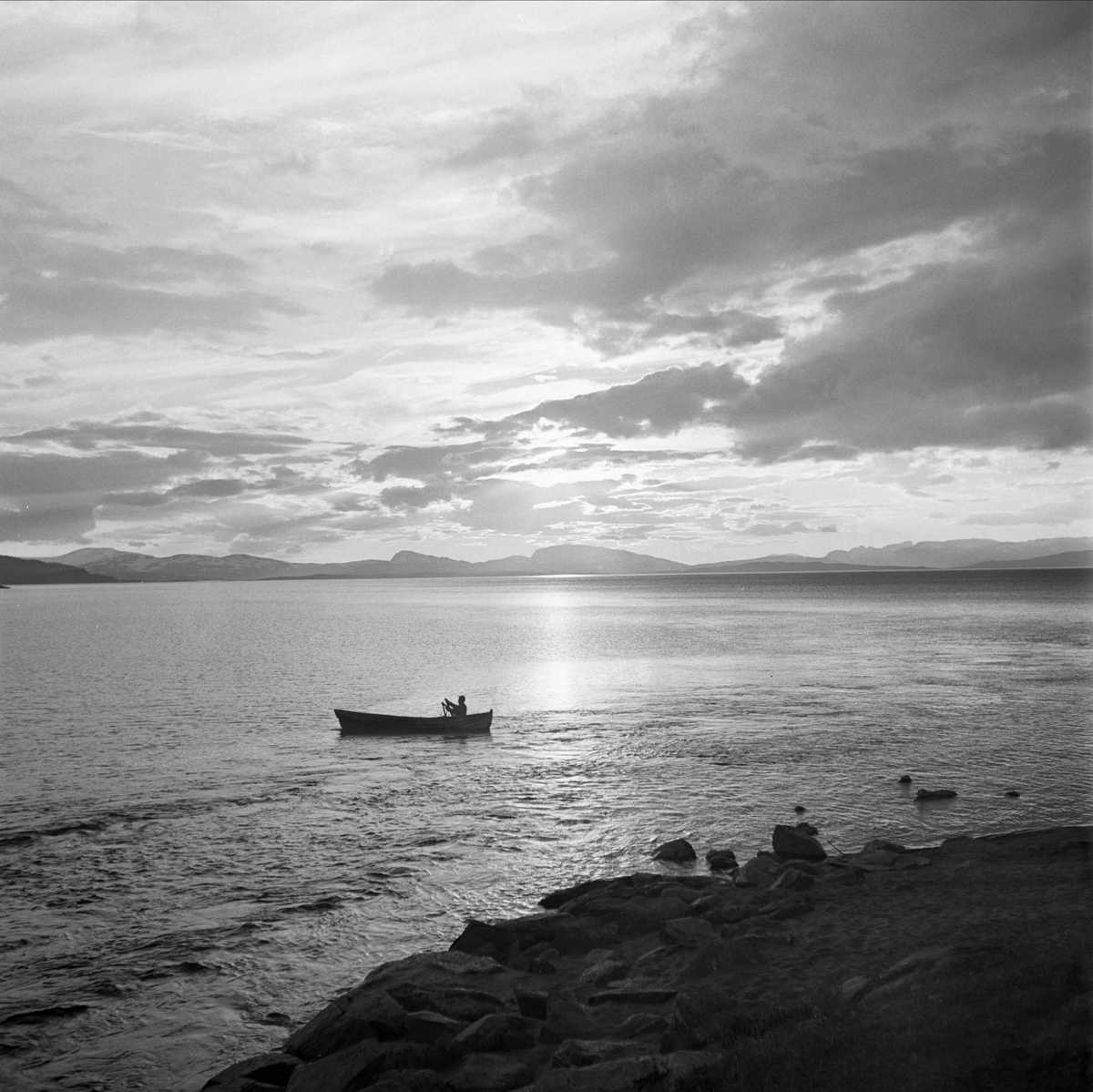 Dag Hammarskjöld på fjällvandring på Sulitelma. Vy över vattnet, med fjällen i bakgrunden. En man ror en båt.