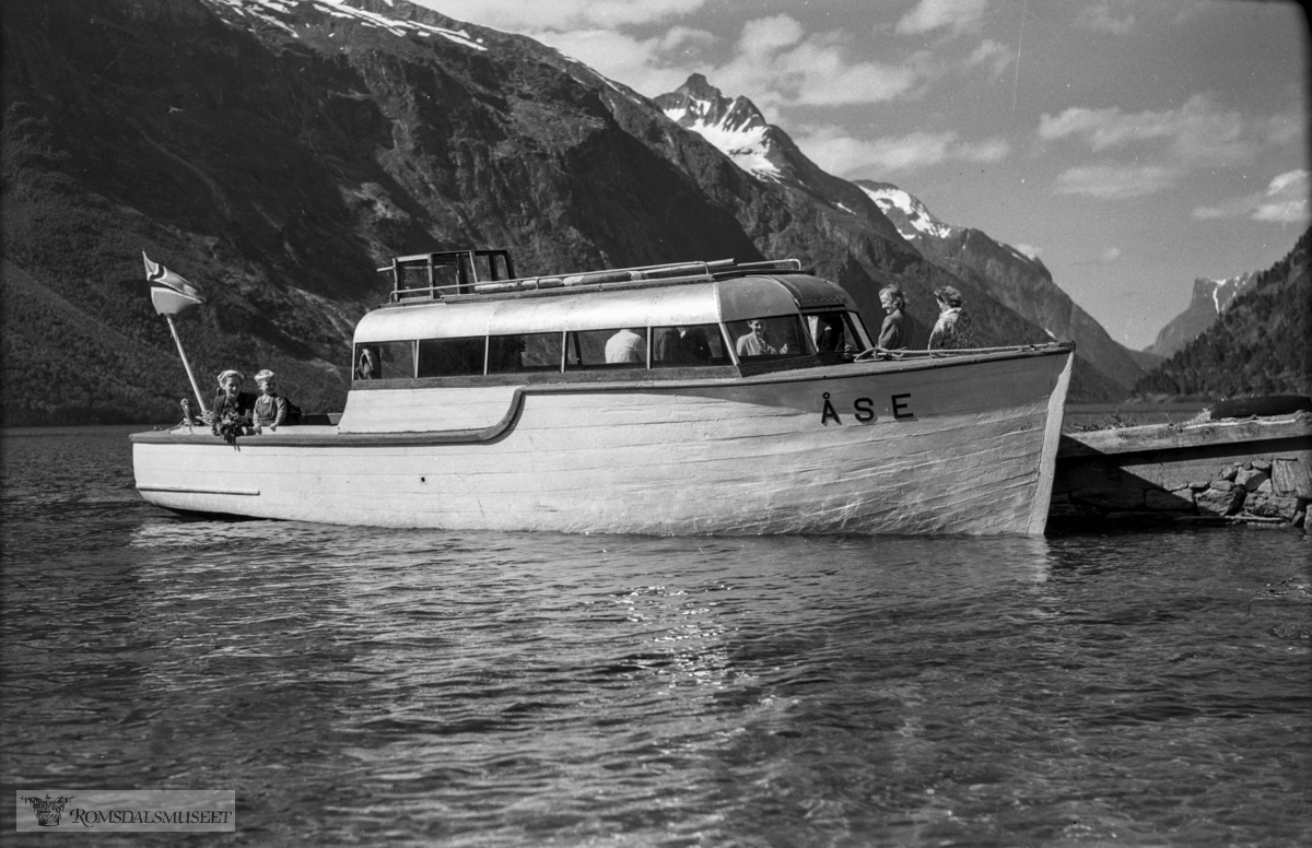 "Fra Eikesdalen, pinsen 1954".Peder Vike (1876-1966) sin private båt "Åse". Båten låg til vanleg ved "Tønna" på Øverås når han ikkje var i bruk..Han frakta folk privat på Eikesdalsvatnet frå ca. 1945 til slutten på 1950-talet.