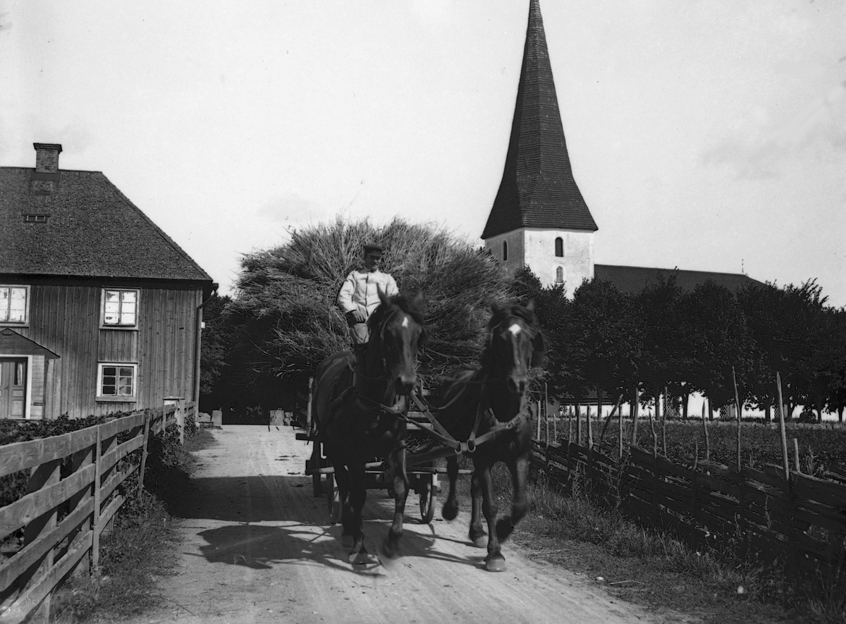 Ögonblick från Viby omkring 1905, där ett imponerande lass hämtas hem till någon av byns lantbruk. Till vänster ligger Källgården med ortens handelsbod, som vid tiden upplysningsvis drevs av bildens fotograf, Carl Sundström. Till höger församlingens kyrka som stått på platsen sedan 1100-talet.