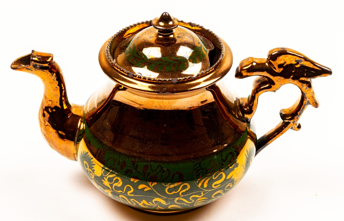 Tekanna, keramik, glasyr övervägande i starkt kopparglänsande färg, med bård av grönaktiga blomster nertill.
