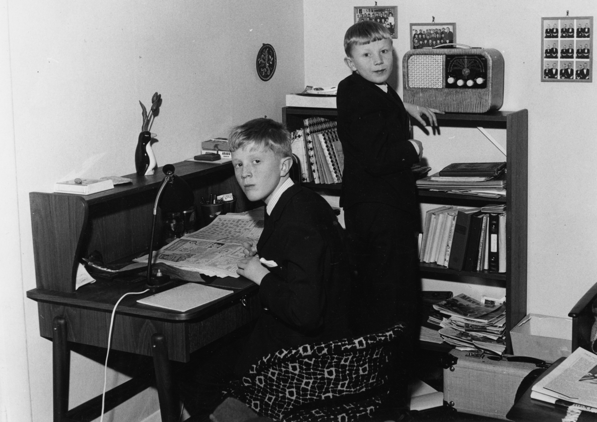 Foto av to dresskledde gutter på et typisk norsk gutteværelse fra 50/60-tallet. Den ene gutten står ved siden av en radio, den andre sitter ved en skrivepult, han har en avis foran seg oppslått på "lørdags kroken" med kryssord og liknende oppgaver. I hyllene ligger tegneserier.