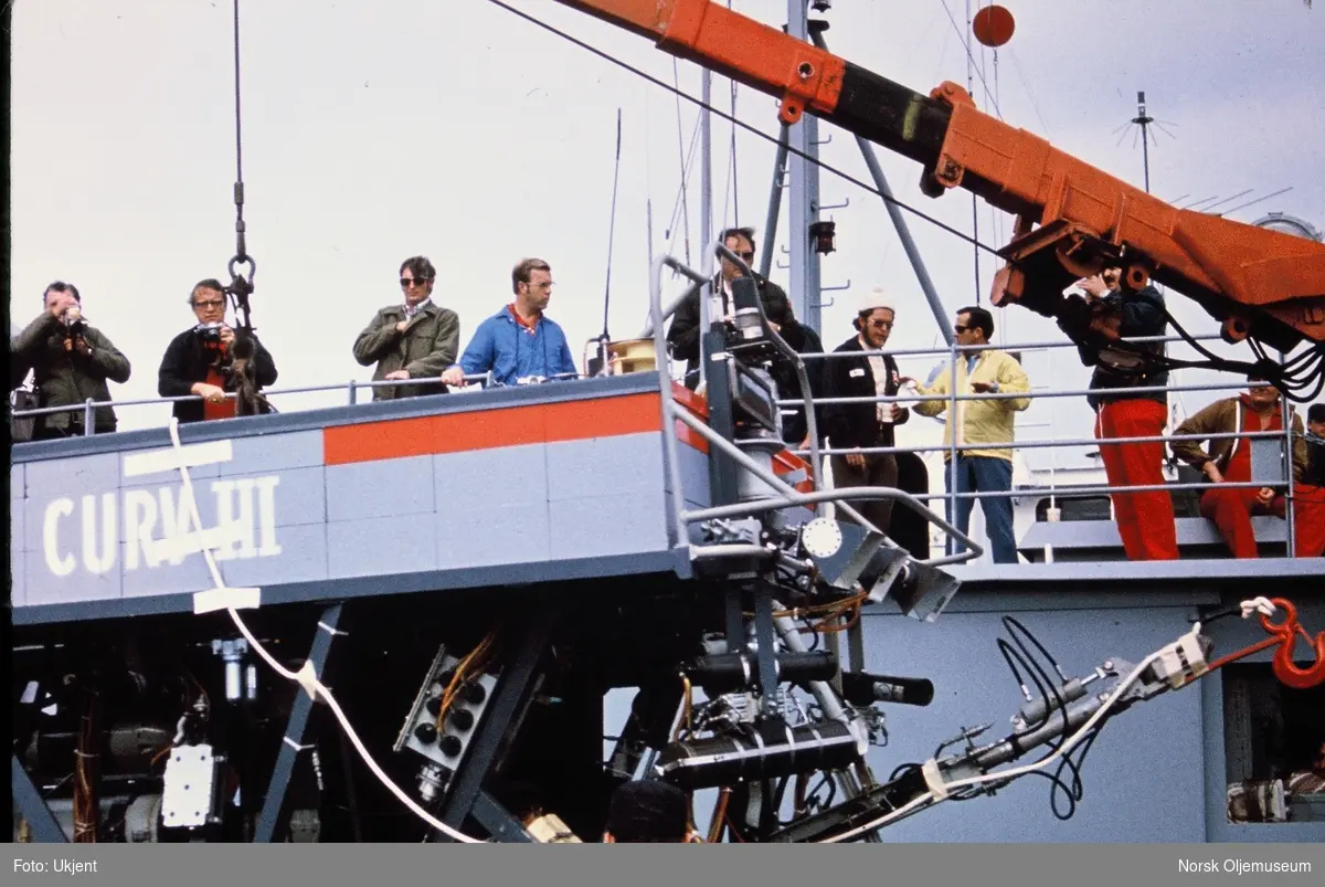 Et stort undervannsfartøy ved navn "CURV-III" i en wire utenfor et skipsdekk. På dekk står flere menn og følger med. Noen filmer og fotograferer og en har en flaske sprudlevann.