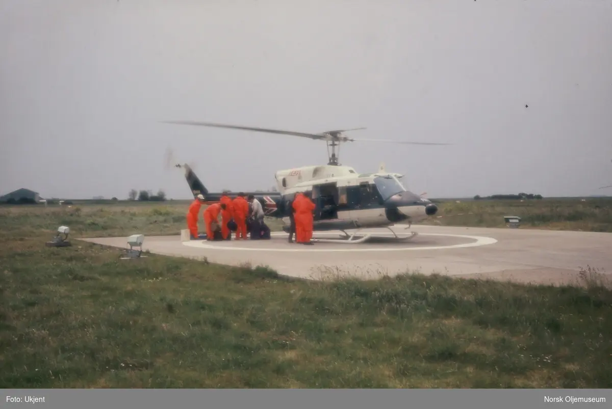 Arbeidere gjør seg klare for transport med et britisk BEA helikopter. Alle er kledd i orange arbeidsdresser.