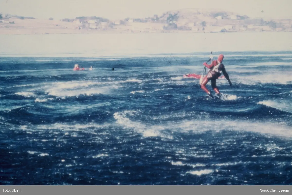 En livredder fires ned fra et helikopter til menn i overlevelsesdrakter i sjøen. På bilde 2 fester han et belte rundt mannen i sjøen og på det siste bildet heises de opp av sjøen.