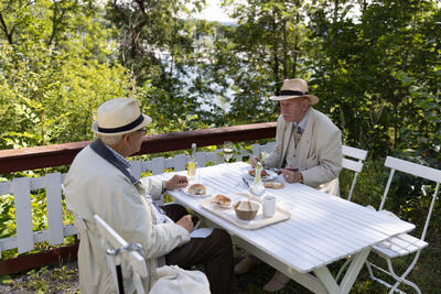 To menn sitter ved et bord og spiser lunsj i grønne omgivelser. 