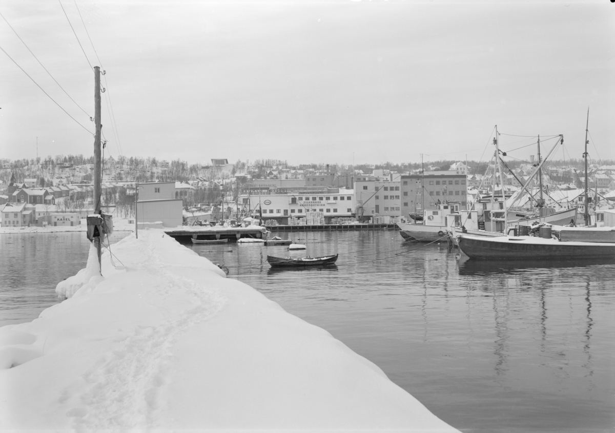 Landskapsfoto av havn i Tromsø om vinteren. Fiskebåter og en robåt ligger i havnen utenfor Tromsø Fryseri / Fiskernes Agnforsyning. Macks Bryggeri skimtes bak.