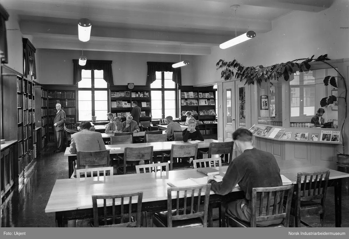 Mennesker sittende i bibliotekssalen / lesesalen på Rjukan Bibliotek. Kvinnelig biblotekar arbeider på bibliotekets kontor.