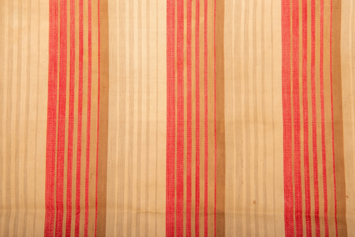 Bolster, bomull, satengvev.
Rødt, brunt, beige i brede striper. Hver inndelt i smale striper.
En kortside delvis sydd igjen for hånd med sort tråd. En rift sydd igjen.