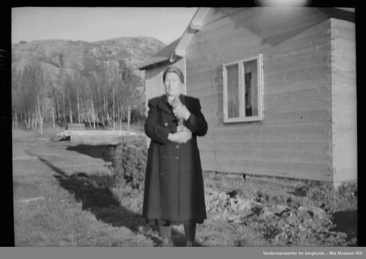 Kvinne med kattunge utenfor et hus med byggemateriale og fjell i bakgrunnen.