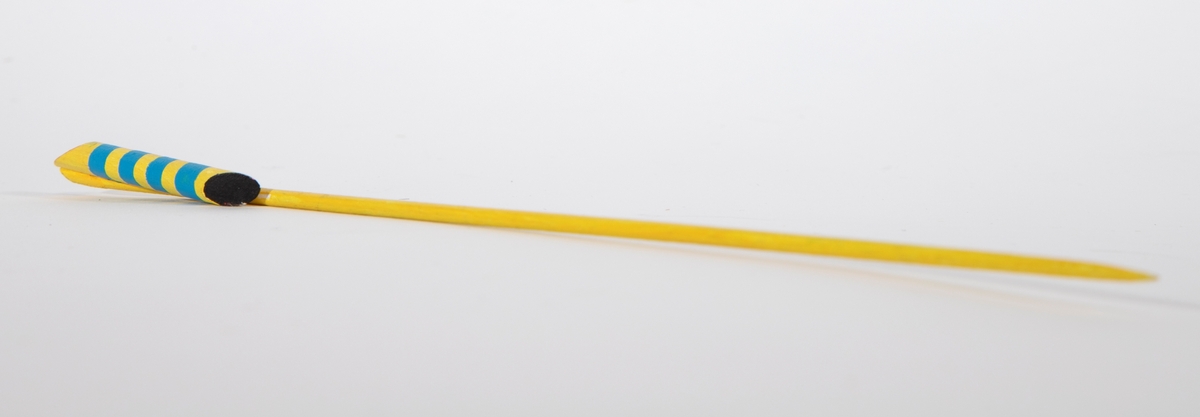 Brosje bestående av en lang gulmalt trepinne med spiss ende. Øverst, parallelt med pinnen er det limt på en bit av en tykkere trepinne som er malt gul med blå striper. De skrå endene er malt i henholdsvis rød og svart.