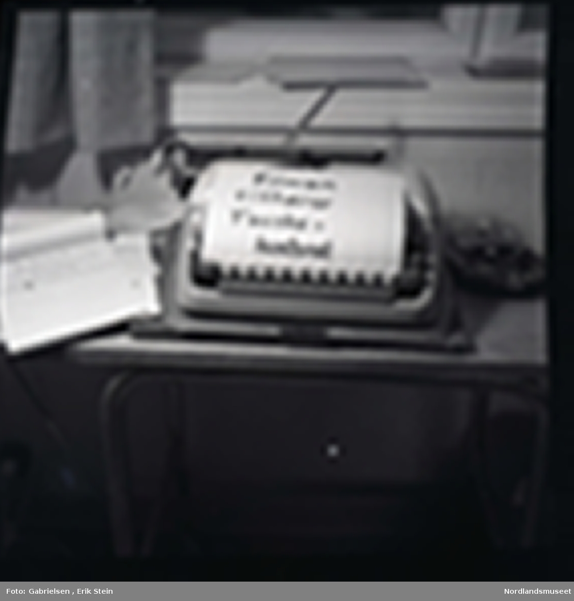 Fotografi av en gammel skrivemaskin 
som står på en pult inne i et kontor under en vindu med noen gardiner som det ligger en lapp på som står filmen tilhører 
Fauskekontoret og det ligger noen papirblokker
vedsiden av skrivemaskinen og man ser noen
mapper som ligger på vinduskarmen i kontoret 
og man ser en ovn under pulten i kontoret