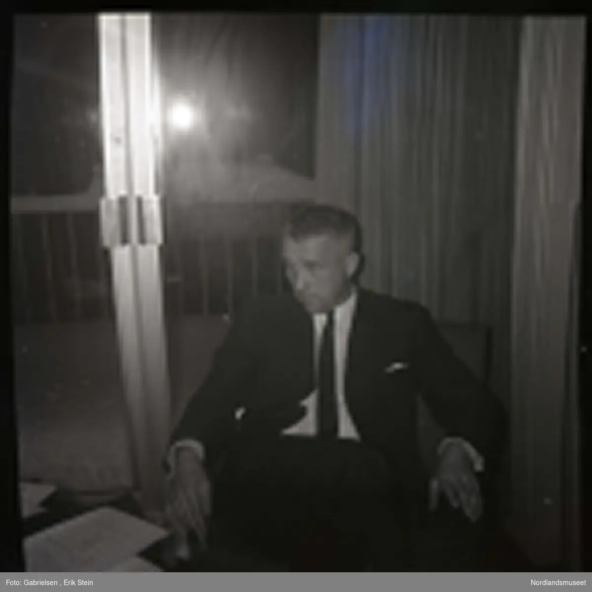 Fotografi av en mann kledd i en dress med slips 
som sitter på en stol foran en dobbeldør til en balkong
i en stue vedsiden av et bord som det ligger noen papirbunker og bøker på og man ser at det har snødd på balkongen ute og man ser et nøkkelhull til dobbeldøren
til balkongen