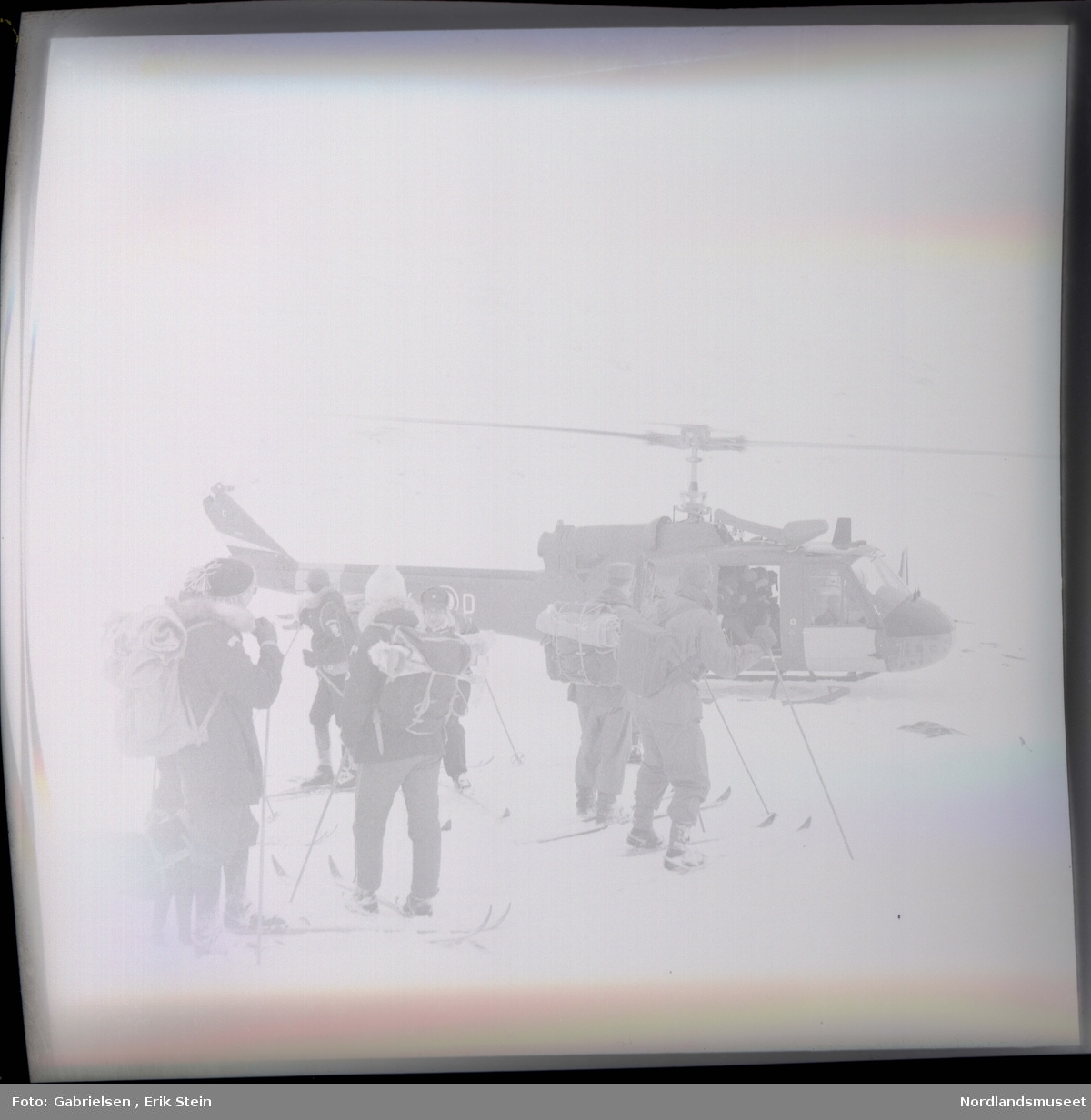 Fotografi av mange menn og kvinner kledd i vinterklær
med sekker på ryggene som står på en snøslette med ski på seg i næreheten av et helikopter som man
ser en mann inni i døråpningen til helikopteret 
som man ser står på snøsletten ved et berg