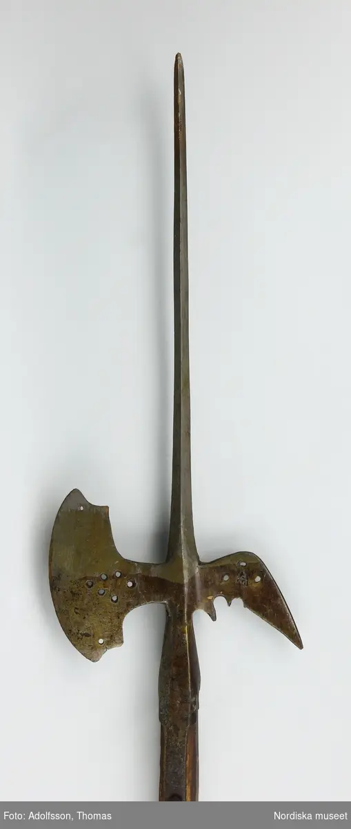 Hillebard, med  originalskaft, oktogonal hyvlad ek. Hillbarden tillhör en typ av svensktillverkade vapen som under 1500-talets mitt och senare hälft utbildades ur tyska förebilder. Den har lång smal stötklinga, med fyrkantigt tvärsnitt. Både hake och yxblad är genombrutna av små, runda hål vilka bildar ornament. Den är  tillverkad i Arboga och i detta fall känner vi tillverkaren vid namn genom den stämpel som återfinns på vapnet. Det är Philpus Osterreich eller Philpus hillebardsmed som tillverkat det och varverksam vid Arbogafaktoriet mellan åren 1567 till 1609. 
/Johan Åkerlund 2006-03-16.
