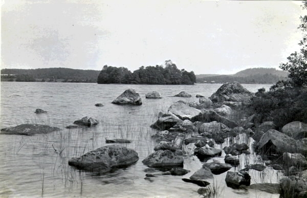 Stenig strandkant i Sjönevadssjön med vy mot ön med lämningarna av Sjönevadsborg.