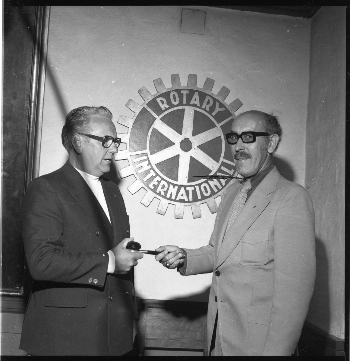Två män med en ordförandeklubba emellan sig. De står framför en skylt med logo för Rotary.