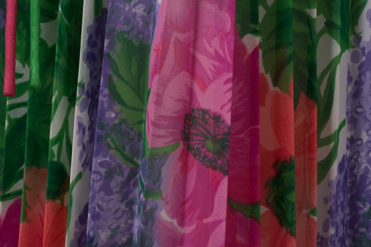 Lång skjortblusklänning i polyester med permanentplisserad kjol. 
Blommigt mönster i vitt, rosa, rött, grönt och lila. På framsidan finns tre klädda knappar och ett blixtlås. De långa ärmarna är rynkade vid handleden och knyts ihop med band. Till klänningen hör ett löst knytskärp av ett rosa och ett grönt stoppat band. 
Blixtlåset fram är fastsatt vid ett senare tillfälle samtidigt som ett knapphål och en knapp sytts dit mitt fram. 
Kjolen är fodrad med ett vitt nylontyg.

På insidans etiketter framgår att klänningen har storlek 42 och är tillverkad i Sverige för "collection philip". På tvättetiketten är tvättrådet 40 grader, ingen torktumling, låg värme på strykjärnet samt kemtvätt "P".