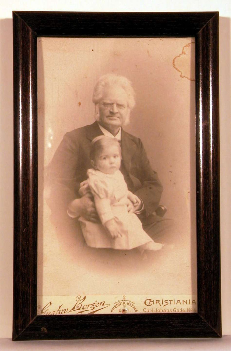 Atelierfortografi; portrettfoto av B.B. sittende med barnebarn på fanget. 