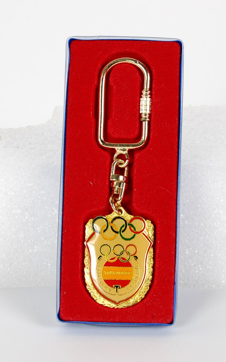 Gullfarget nøkkelring med merke med flerfarget logo for det som ser ut til å være det spanske taekwondo-forbundet. De olympiske ringene inngår i logoen. Det er også gravert inn en taekwondo-logo på baksiden. Nøkkelringen ligger i en rød og blå eske av plast.