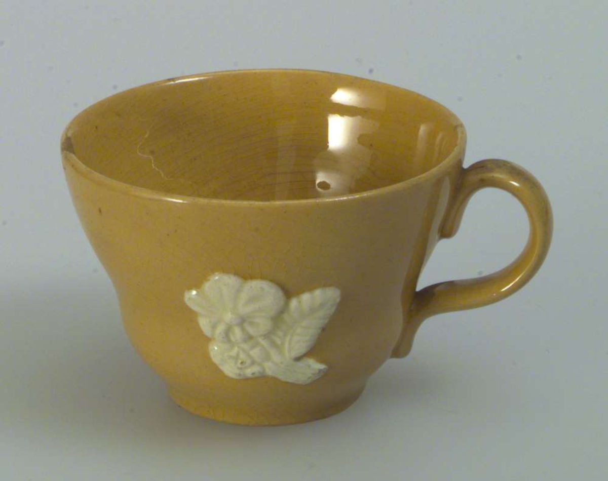 Gul kaffe-/tekopp med skål. Koppen er dekorert med en blomst med blad. Dekoren er hvit. Skålen er uten dekor. Det er hakk i både koppen og skålen.
Det er ingen produksjonsmerker på kopp eller skål.