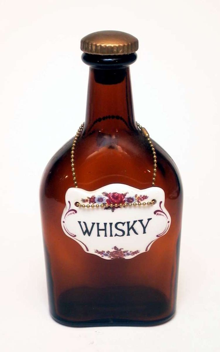 Flat brennevinsflaske i brunt glass med kork. Rundt halsen på flasken henger et skilt i lenke. Det er av hvitt glass, dekorert med blomster og er påskrevet 'Whisky'.