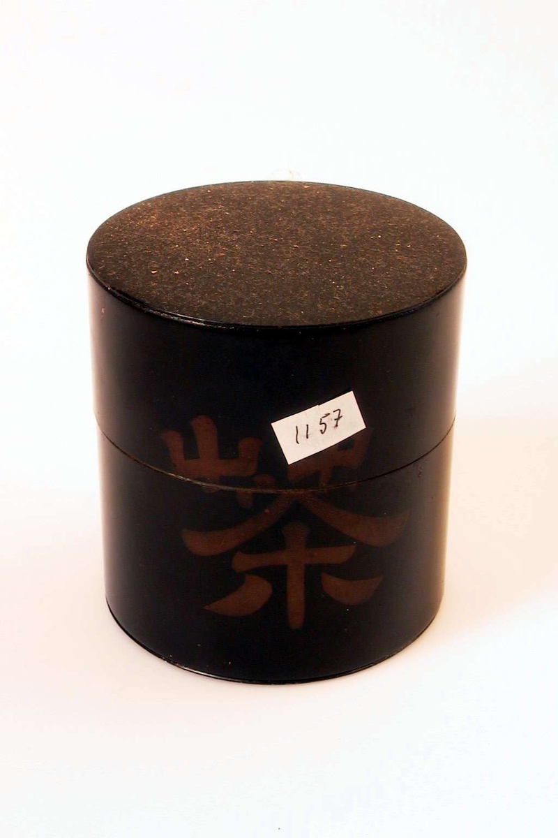 Boks av metall. Boksen er sort og dekorert med et  kinesisk skrifttegn. Den har et stempel i form av et hode. Boksen har to lokk.