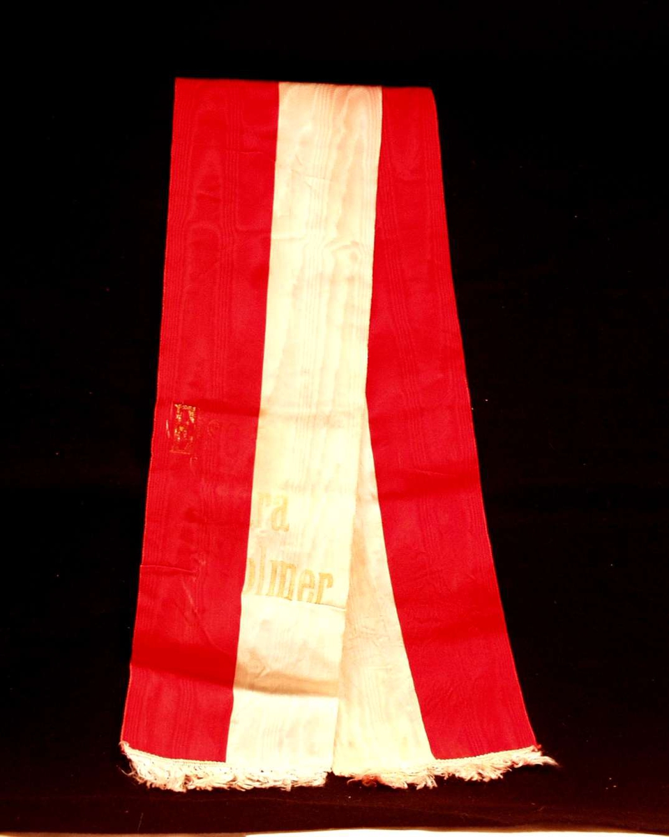 Bånd i rød og hvit silke. Båndet er delt i to. På båndet står det: Else Clara Volmer. De forgylte bokstavene er delvis avslitt.