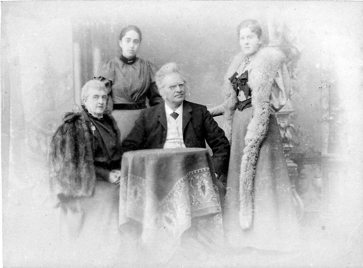 Atelierfotografi; bakerst står to jevngamle unge damer, den ene med lang pelsstola hengende ned langs med skjørtet og en stor mørk sløyfe bundet midt for brystet; den andre unge dame mer enkelt kledd i lys lang kjole med mørkt bånd rundt taljen og halsen isatt enkel brosje. Inn til et lite rundt bord sitter en dame med pelskape til høyre, og i midten en mann med henderne i siden, nesebriller, mørk jakke og slips, hvit skjorte og vest. Teppebakgrunnen viser antikke bygningsfragmenter. De tre damene ser direkte på betrakteren, mens mannen ser skrått til venstre.
