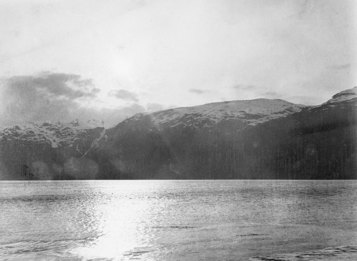 Fjordlandskap i Hardanger.  Fotografiet er tatt over en fjord i motlys, slik at solstrålene speiler seg i vannflata.  På motsatt side av fjorden er det høye fjell med snø på toppene. 