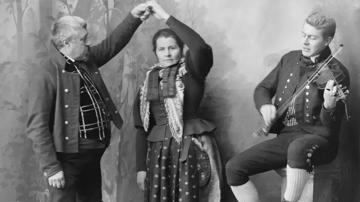Dette er et gruppebilde der mann og kvinne ikledd Rørosdraktdanser pols. Felespiller sitter ved siden av.