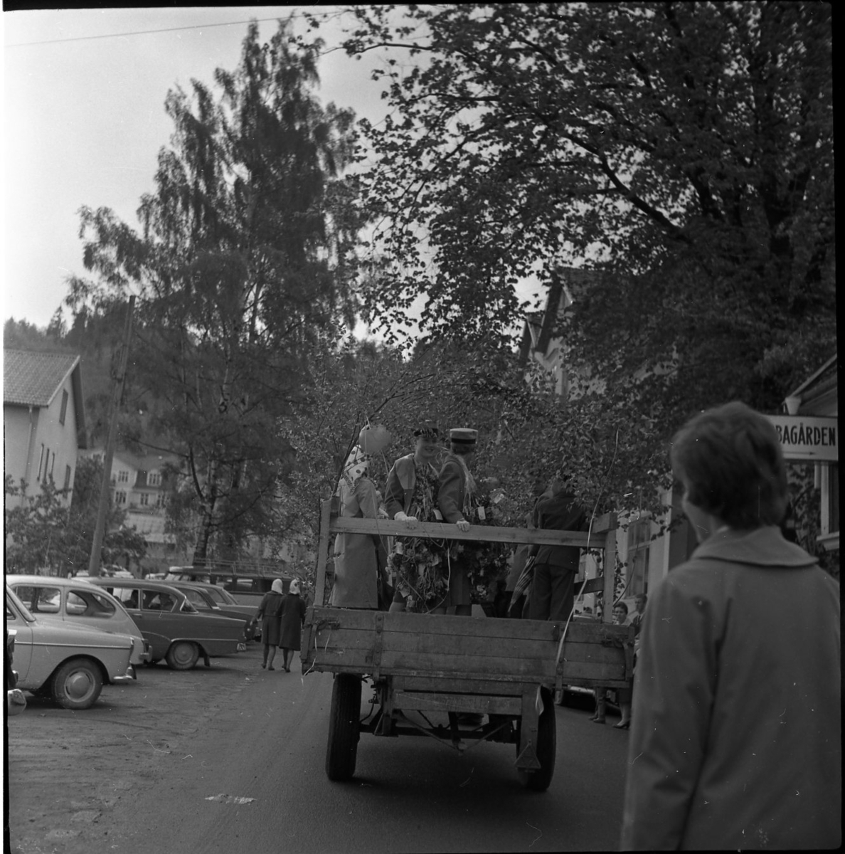 Realexamen 1961. Bilden föreställer ett traktorsläp fotograferad bakifrån. Den är klädd med björkris och ballonger. På släpet står glada realare. Bredvid lsatbilen står personer och tittar.